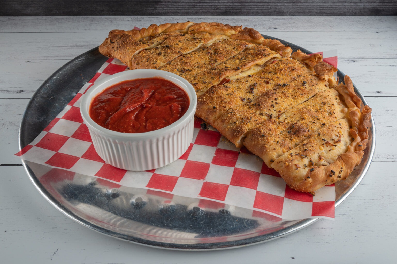 Golden-brown calzone served on a metal tray with red checkered paper, accompanied by a small bowl of marinara sauce. Perfect for sharing.